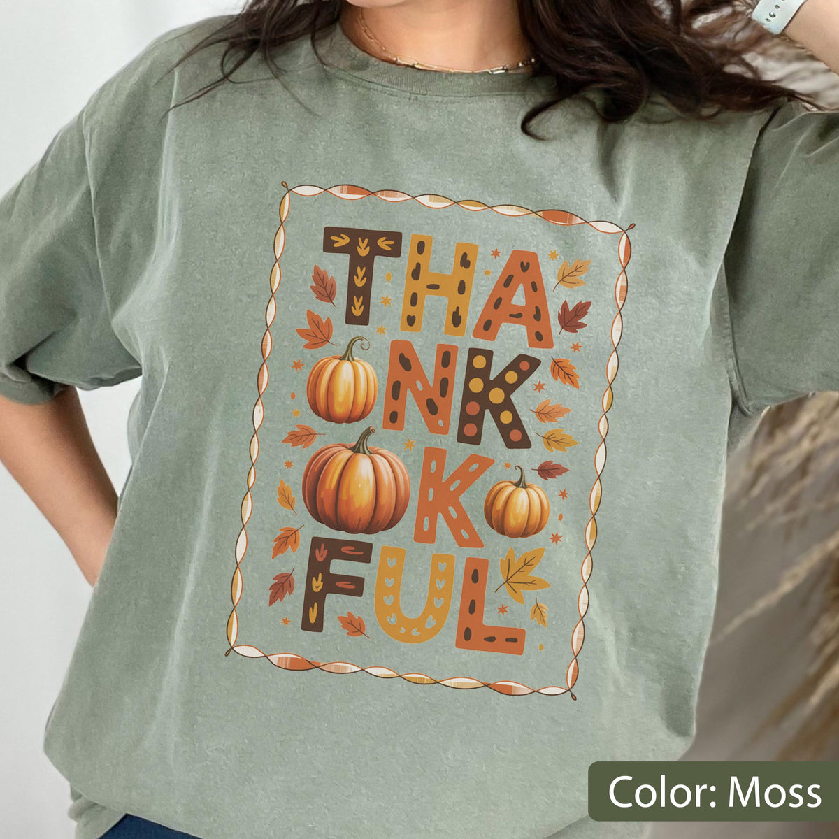 May include: Moss green t-shirt with the word "THANKFUL" in orange and brown letters, decorated with pumpkins and fall leaves. The design is framed with a brown border. The shirt is a relaxed fit.