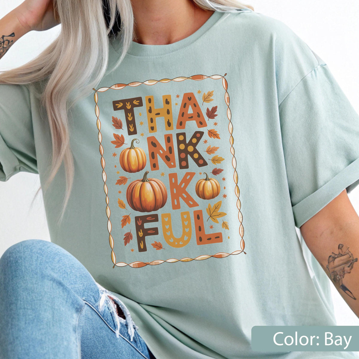 May include: A light blue t-shirt with the word "THANKFUL" in a decorative frame. The word is spelled out with orange and brown letters, pumpkins, and fall leaves. The shirt is a solid color and has short sleeves.