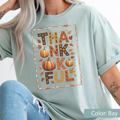 May include: A light blue t-shirt with the word "THANKFUL" in a decorative frame. The word is spelled out with orange and brown letters, pumpkins, and fall leaves. The shirt is a solid color and has short sleeves.