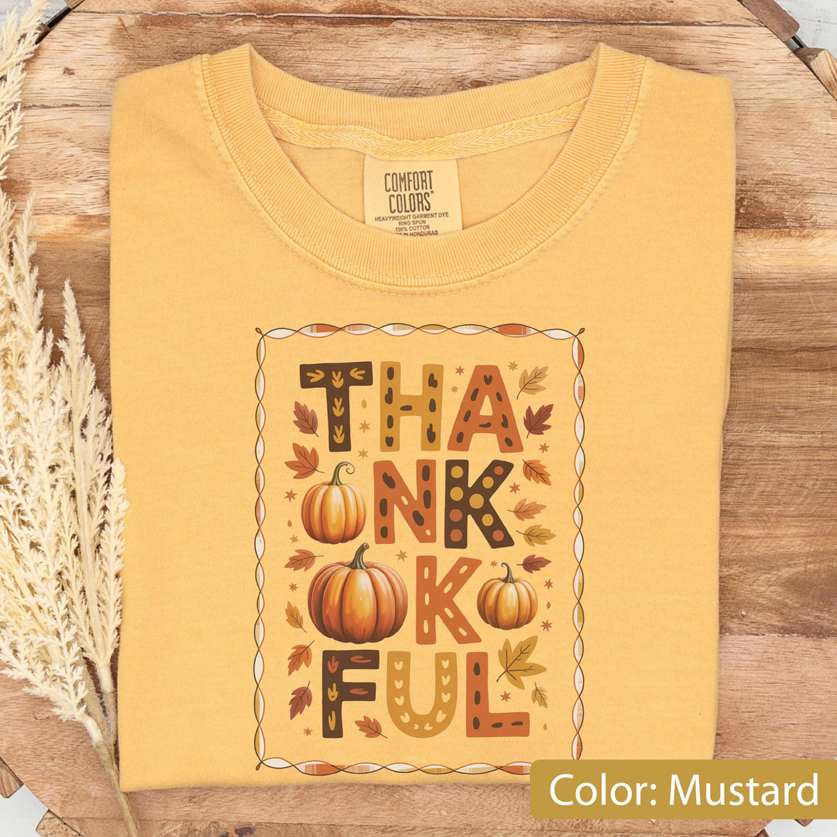 May include: Mustard-colored t-shirt with the word "THANKFUL" in a decorative fall-themed design. The design includes pumpkins and autumn leaves. The shirt is made of a soft material and has a crew neck.
