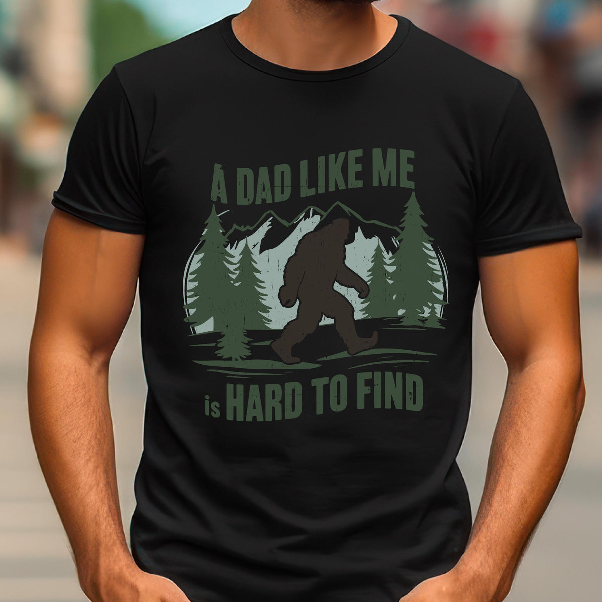 May include: A black short-sleeved t-shirt featuring a graphic print of a brown Bigfoot silhouette walking through a forest with mountains in the background. The text reads A DAD LIKE ME is HARD TO FIND in green and white lettering on the front.