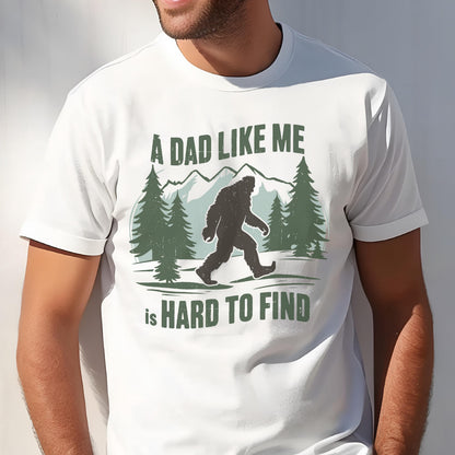 May include: A white short-sleeved t-shirt featuring a graphic print of a black Bigfoot silhouette walking through a forest with mountains in the background. The text reads A DAD LIKE ME is HARD TO FIND in dark green capital letters.