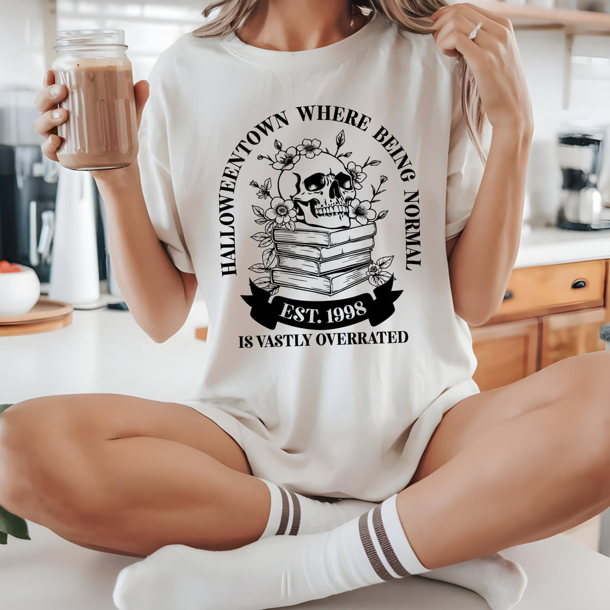 May include: Cream-colored t-shirt with a black graphic featuring a skull atop a stack of books, surrounded by flowers. The text reads "Halloweentown Where Being Normal Is Vastly Overrated" and "Est. 1998".