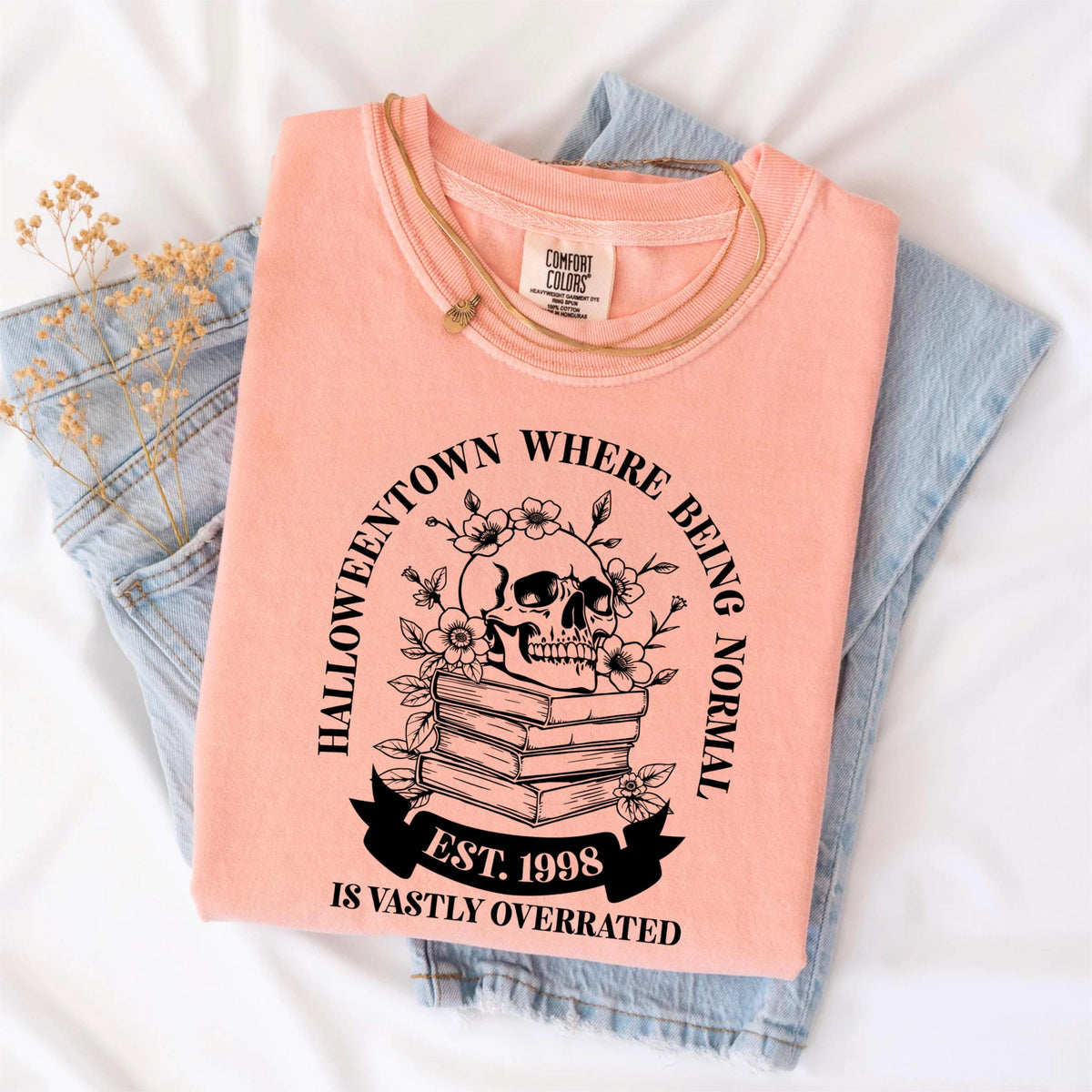 May include: A peach-colored t-shirt with a black graphic featuring a skull, flowers, and the text "Halloweentown Where Being Normal Is Vastly Overrated, Est. 1998." The shirt is folded on top of blue jeans.