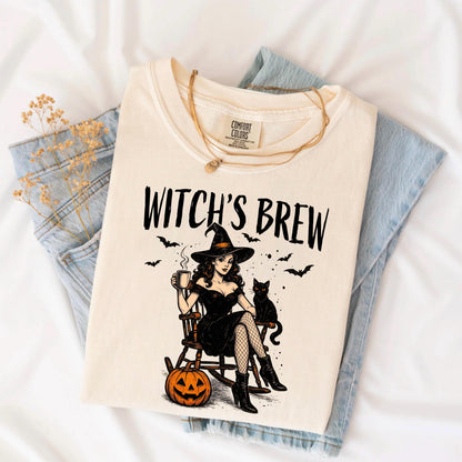 May include: A cream-colored t-shirt featuring a graphic print of a person in a black dress and witch hat sitting in a rocking chair with a black cat, a mug, and a jack-o'-lantern. The text WITCH'S BREW is printed in black above the illustration.