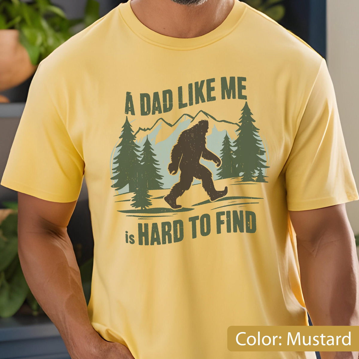 May include: Mustard yellow t-shirt with a Bigfoot graphic and the text "A DAD LIKE ME is HARD TO FIND". The design includes trees and mountains, creating an outdoor theme.