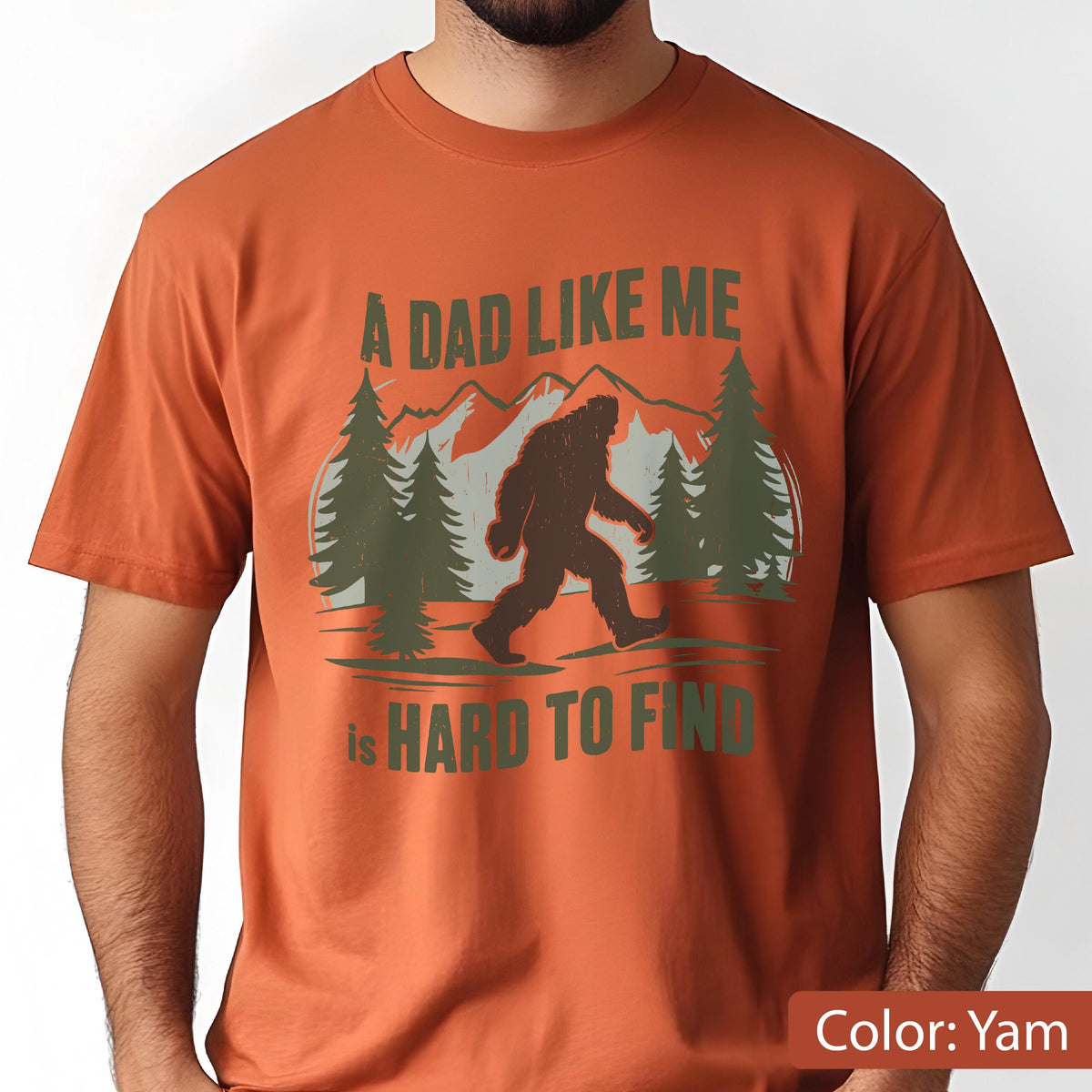 May include: A yam-colored t-shirt featuring a graphic of Bigfoot walking in front of a mountain and pine tree scene. The text reads "A DAD LIKE ME is HARD TO FIND".