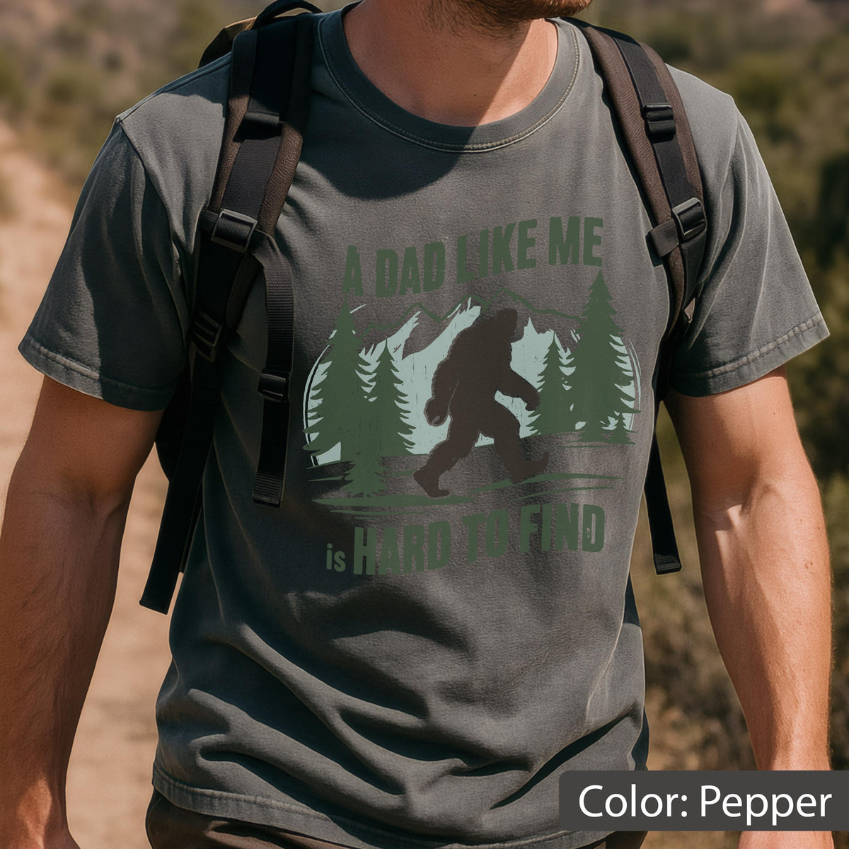 May include: Pepper-colored t-shirt featuring a Bigfoot graphic with the text "A Dad Like Me is Hard to Find." The design includes a silhouette of Bigfoot walking in front of mountains and trees. The shirt is a crew neck and short-sleeved.