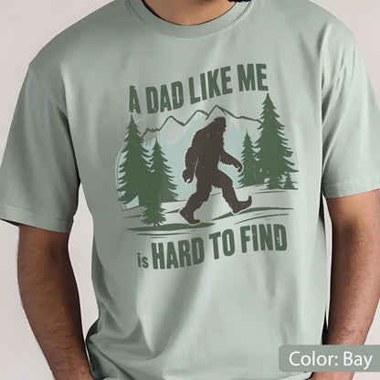 May include: Bay-colored t-shirt featuring a graphic of Bigfoot walking in a forest scene with mountains. The text reads "A DAD LIKE ME is HARD TO FIND" in green lettering. The shirt is a solid color.