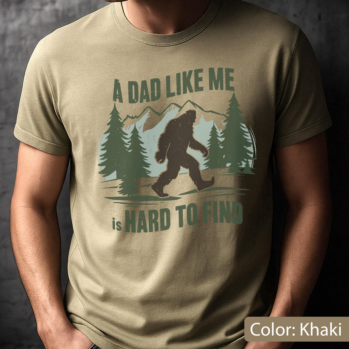 May include: Khaki-colored t-shirt with the text "A DAD LIKE ME is HARD TO FIND" and a graphic of Bigfoot walking in a forest scene. The design includes pine trees and mountains, all in shades of green and brown.
