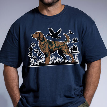 May include: A navy blue t-shirt featuring a graphic print of a hunting dog with a camouflage pattern. The design includes silhouettes of flying ducks, paw prints, and pine trees in white, set against the dark fabric of the shirt.