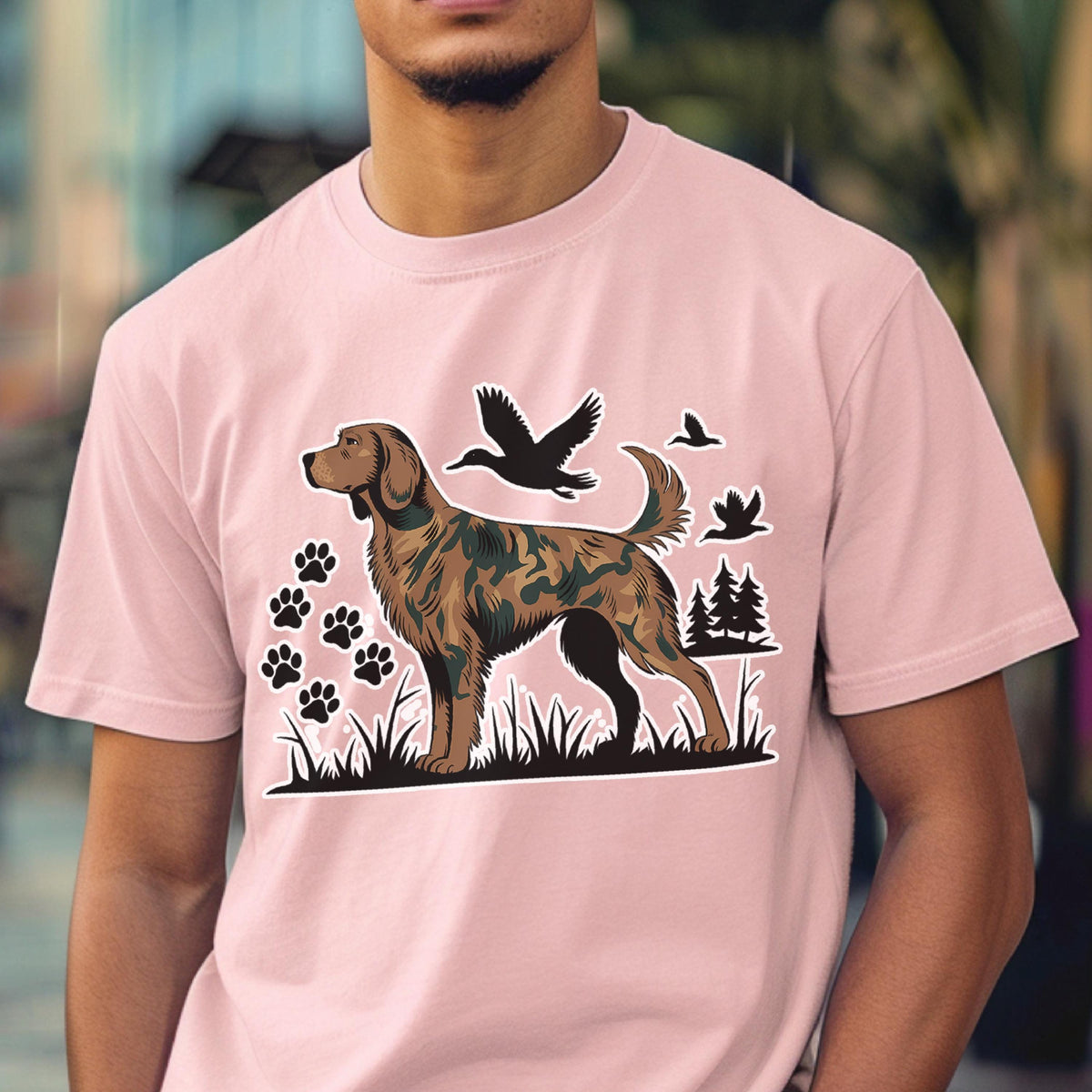 May include: A light pink short-sleeve t-shirt featuring a graphic of a dog with a camouflage pattern. The design includes black silhouettes of flying ducks, pine trees, and paw prints set against a black grassy landscape base.