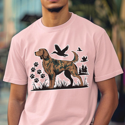 May include: A light pink short-sleeve t-shirt featuring a graphic of a dog with a camouflage pattern. The design includes black silhouettes of flying ducks, pine trees, and paw prints set against a black grassy landscape base.