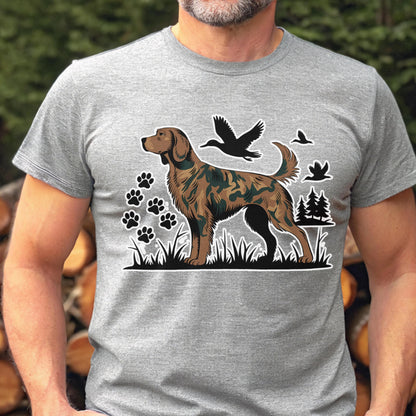 May include: A heather gray short-sleeve t-shirt featuring a graphic print of a brown dog with a camouflage pattern on its body. The design includes black silhouettes of flying ducks, paw prints, and pine trees set against a grassy landscape.