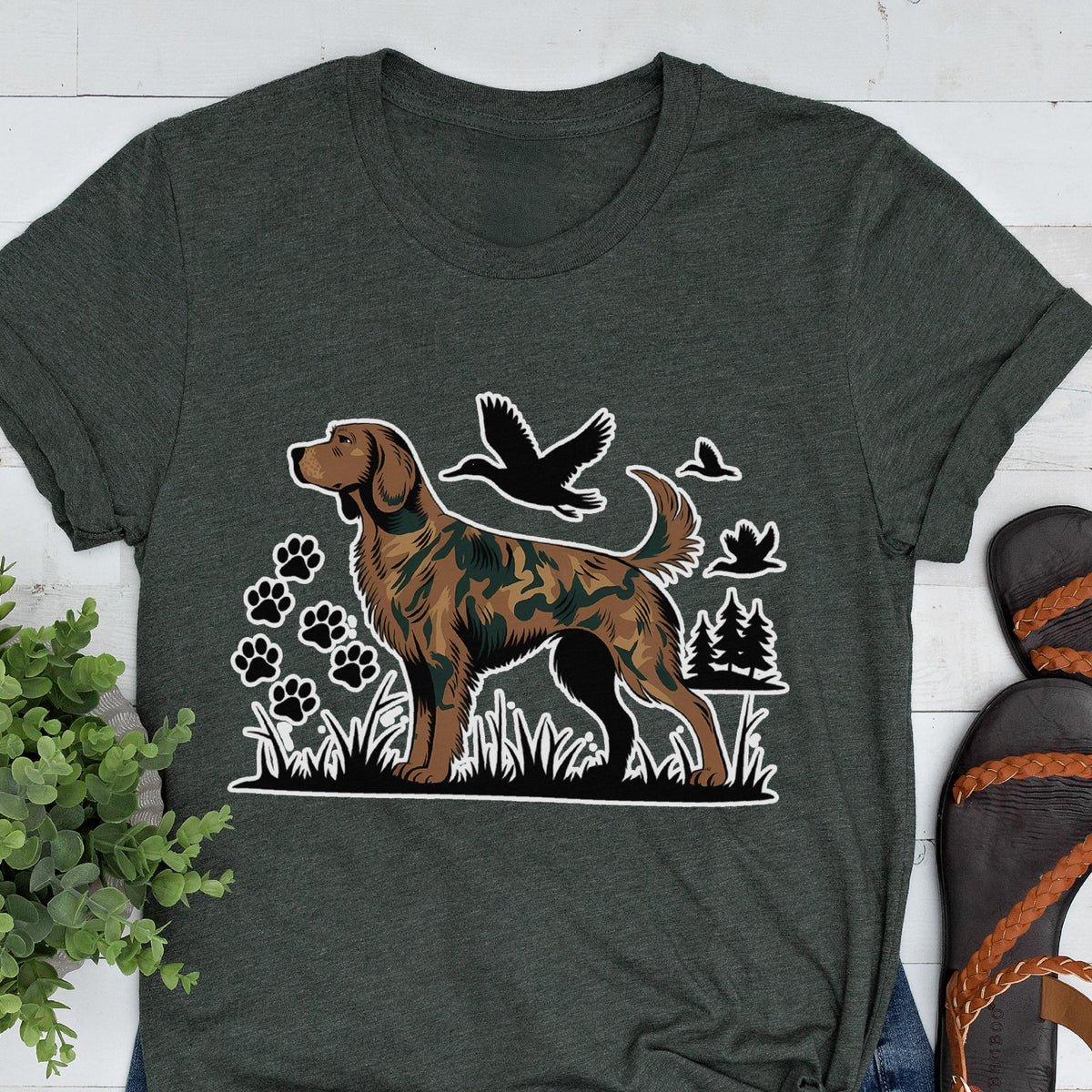 May include: A dark charcoal gray crew neck t-shirt featuring a graphic print of a brown dog with a camouflage pattern. The design includes black silhouettes of flying ducks, paw prints, and pine trees set against a grassy landscape.