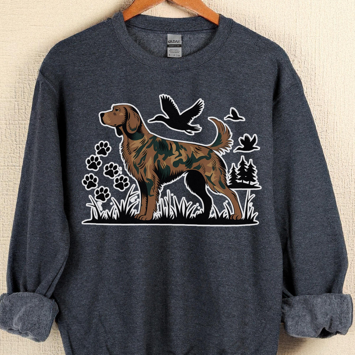 May include: A dark gray sweatshirt featuring a brown dog with a camouflage pattern, standing in grass. Black paw prints, birds, and trees are also in the design. The sweatshirt has rolled-up sleeves and a crew neck.