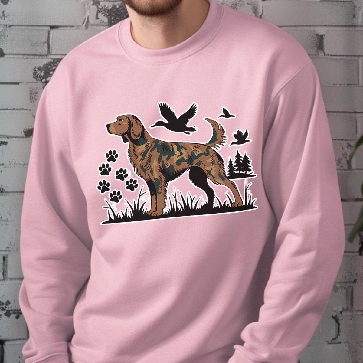 May include: A light pink sweatshirt featuring a brown and green dog illustration with paw prints, trees, and flying birds. The dog has a camouflage pattern. The sweatshirt is made of a soft material.