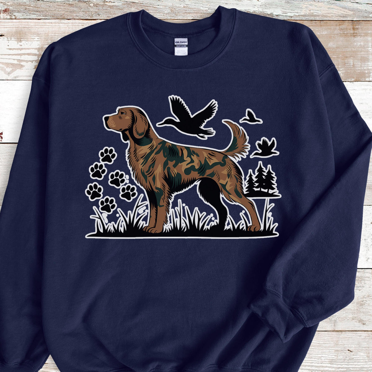 May include: Navy blue sweatshirt featuring a brown dog with a camouflage pattern, standing in grass. The design includes paw prints, birds, and trees. The dog is the central focus, with a white outline. The sweatshirt is a comfortable, casual style.