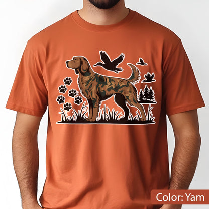 May include: A yam-colored t-shirt featuring a graphic print of a brown camouflage-patterned hunting dog standing in grass. The design includes black silhouettes of flying ducks, a cluster of paw prints, and pine trees in the background.