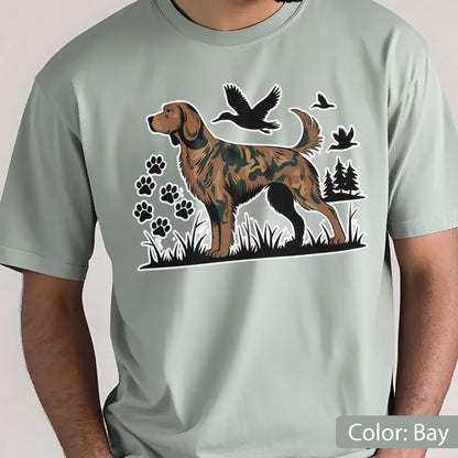 May include: A bay-colored short-sleeve t-shirt featuring a graphic print of a dog with a camouflage pattern on its fur. The design includes black silhouettes of flying ducks, paw prints, and pine trees set against a grassy landscape.
