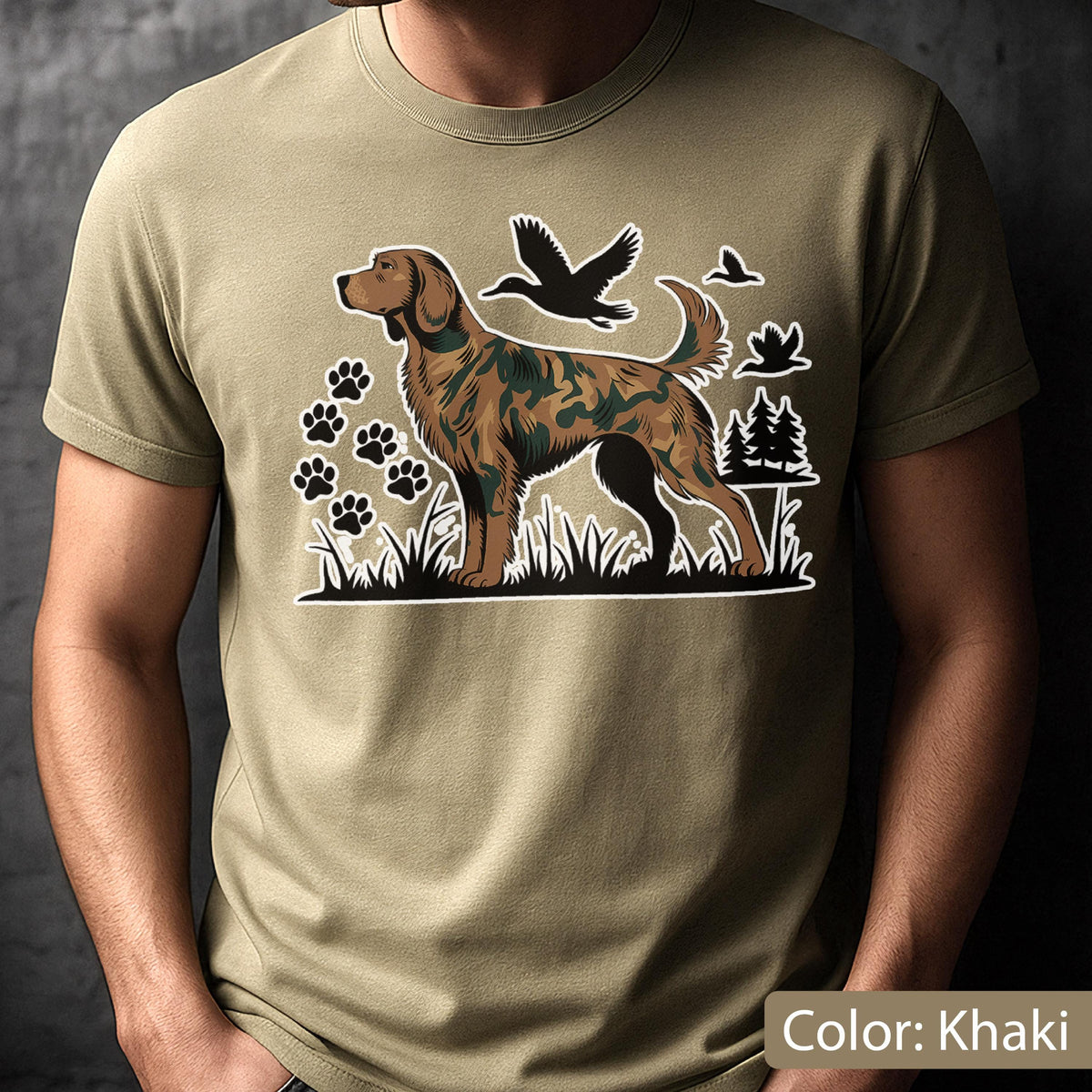 May include: A khaki t-shirt featuring a graphic print of a hunting dog with a camouflage pattern on its fur. The design includes black silhouettes of flying ducks, paw prints, and pine trees set against a grassy landscape background.