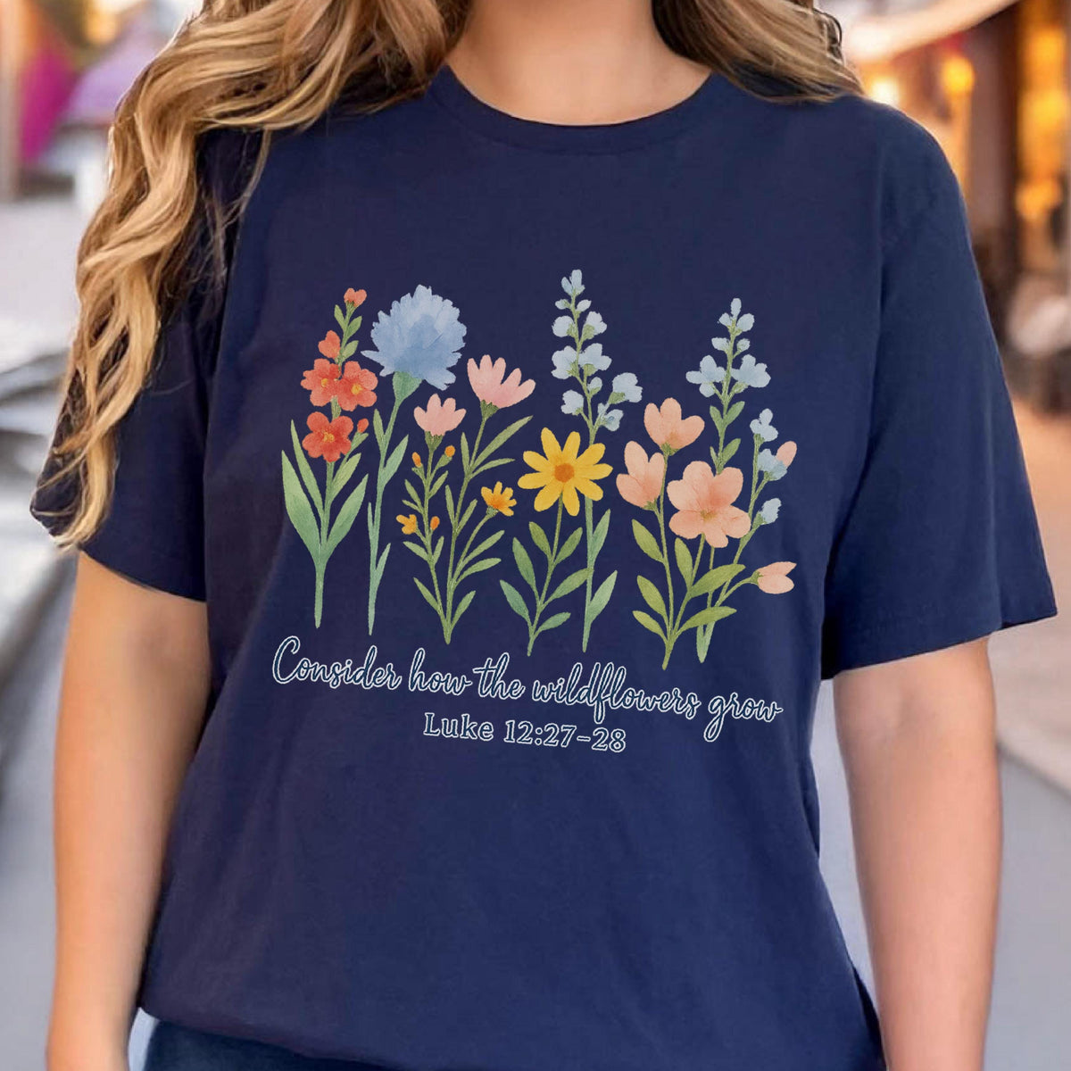 May include: Navy blue t-shirt featuring a colorful wildflower design. The graphic includes orange, blue, pink, and yellow flowers with green stems. The text "Consider how the wildflowers grow" and "Luke 12:27-28" are printed below the flowers.