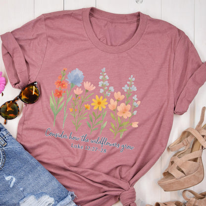 May include: Dusty rose-colored t-shirt featuring a watercolor floral design with orange, blue, yellow, and pink flowers. The text "Consider how the wildflowers grow" and Bible verse Luke 12:27-28 are printed below the flowers.