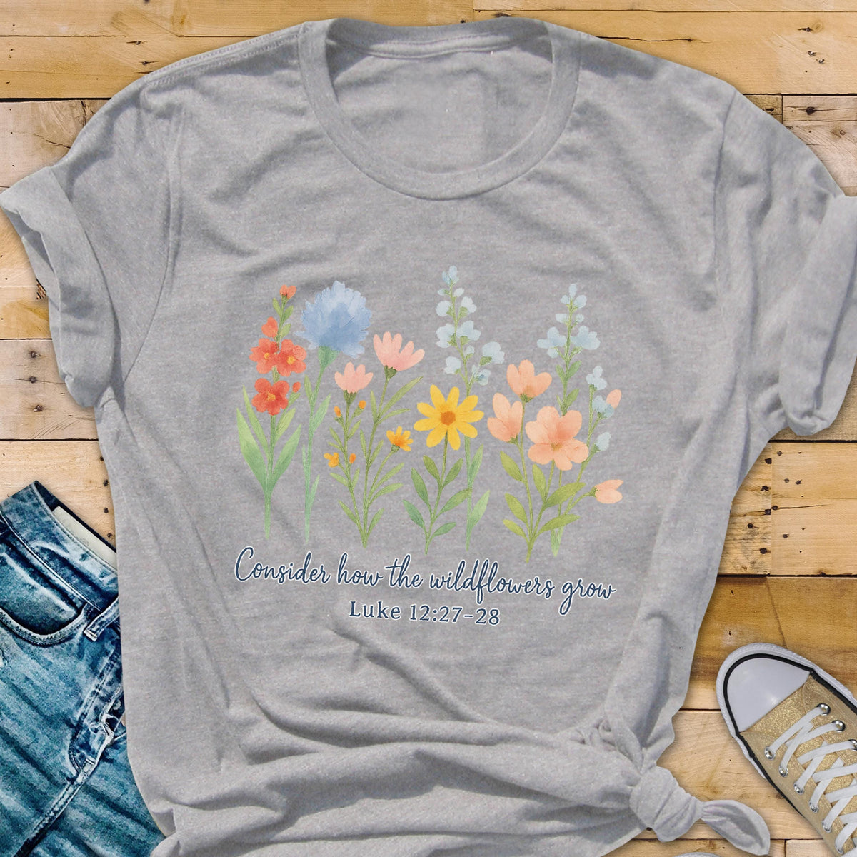 May include: A heather gray t-shirt featuring a watercolor illustration of colorful wildflowers. The text "Consider how the wildflowers grow" and Luke 12:27-28 are printed below the flowers. The shirt is styled with a knot at the bottom.