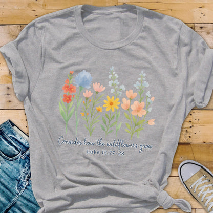 May include: A heather gray t-shirt featuring a watercolor illustration of colorful wildflowers. The text "Consider how the wildflowers grow" and Luke 12:27-28 are printed below the flowers. The shirt is styled with a knot at the bottom.