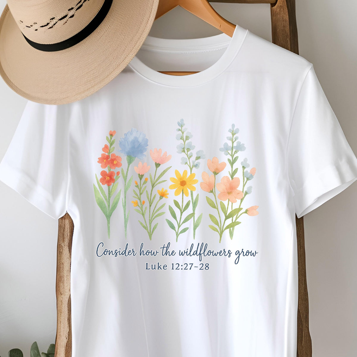 May include: White t-shirt featuring a watercolor floral design with orange, blue, yellow, and peach flowers. The text "Consider how the wildflowers grow" and Luke 12:27-28 are printed in a dark blue script.