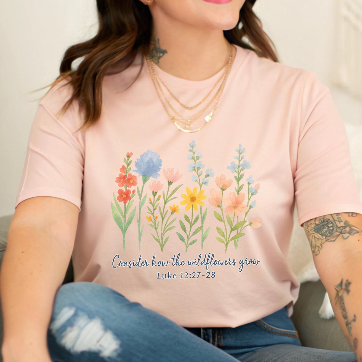 May include: A light pink t-shirt featuring a watercolor floral design with various wildflowers in shades of red, blue, yellow, and pink. The text "Consider how the wildflowers grow" and Luke 12:27-28 are printed below the flowers.