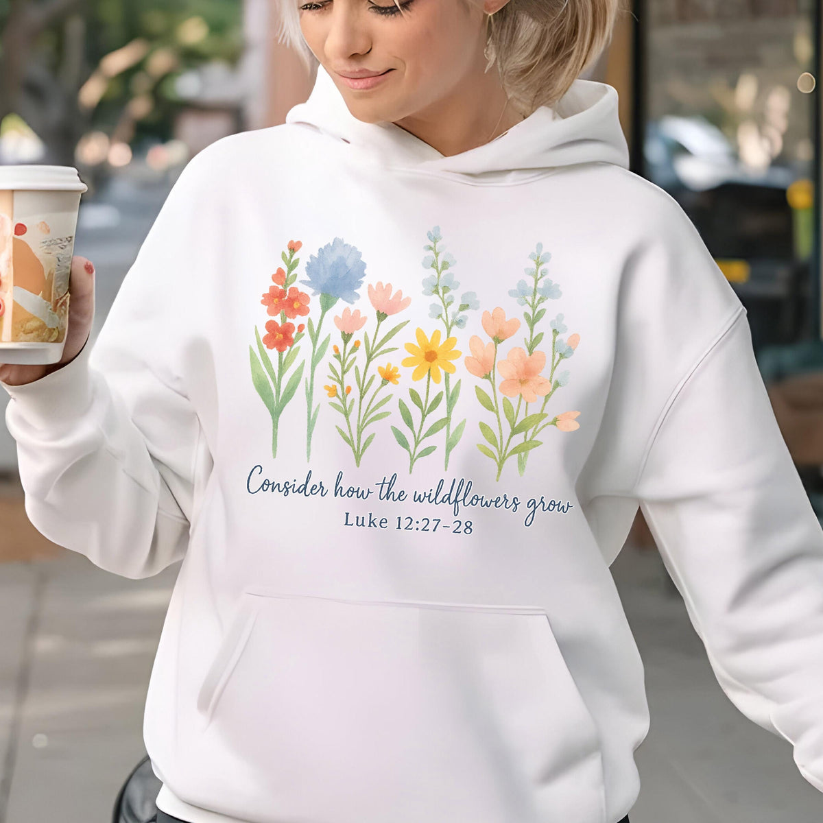 May include: White hoodie with a floral design featuring colorful wildflowers in shades of red, blue, yellow, and peach. The text "Consider how the wildflowers grow" and Luke 12:27-28 are printed in blue script. The hoodie has a front pocket.