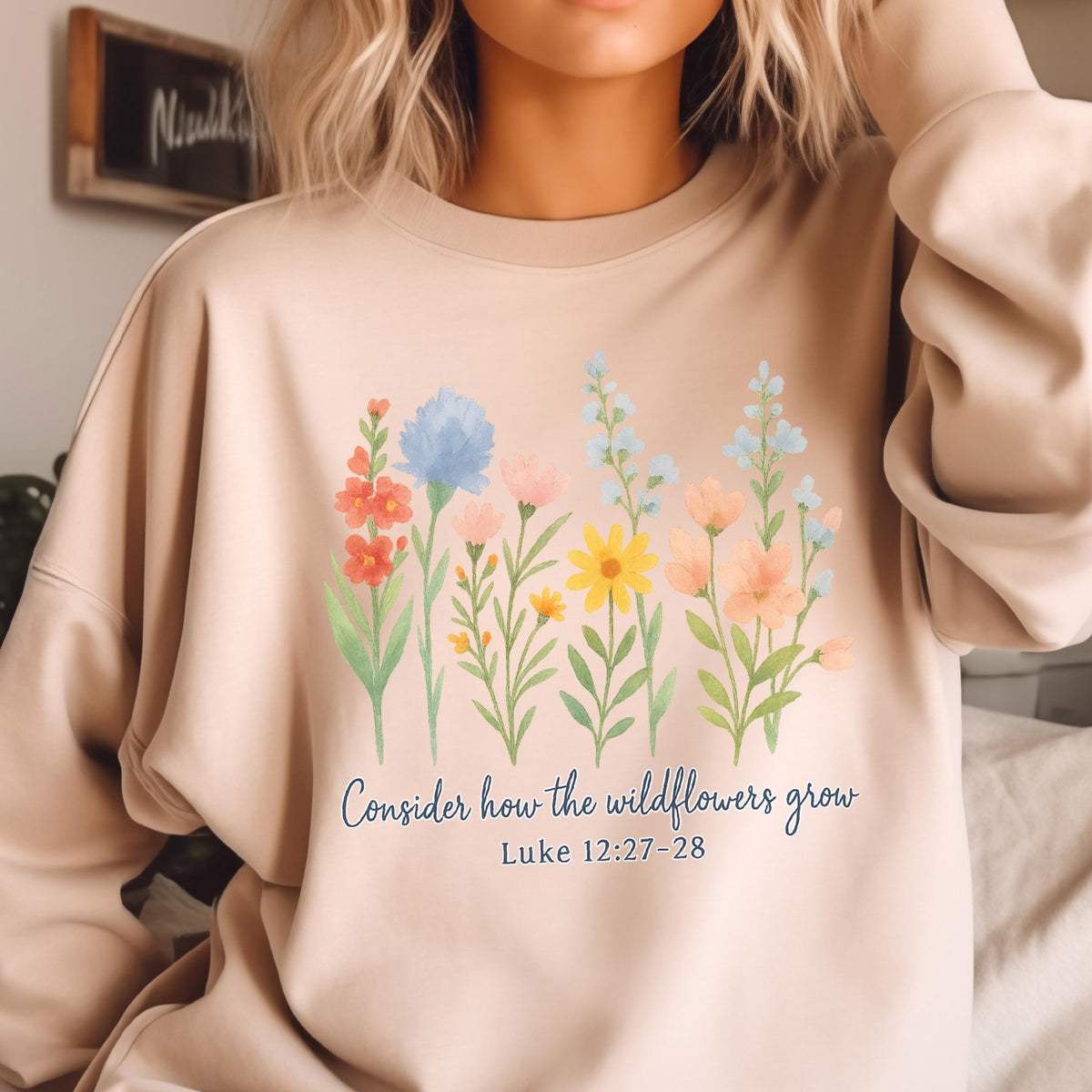 May include: Beige sweatshirt featuring a watercolor floral design with various flowers in shades of red, blue, yellow, and pink. The text "Consider how the wildflowers grow" and Luke 12:27-28 are printed below the flowers.