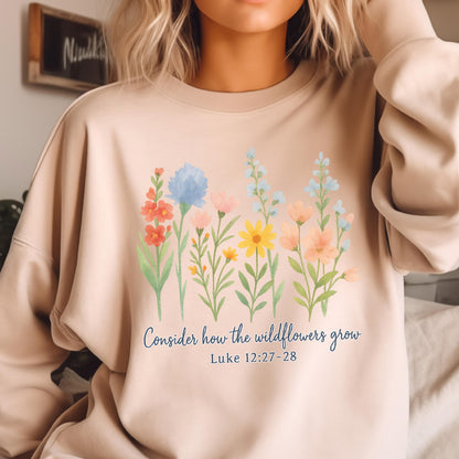 May include: Beige sweatshirt featuring a watercolor floral design with various flowers in shades of red, blue, yellow, and pink. The text "Consider how the wildflowers grow" and Luke 12:27-28 are printed below the flowers.