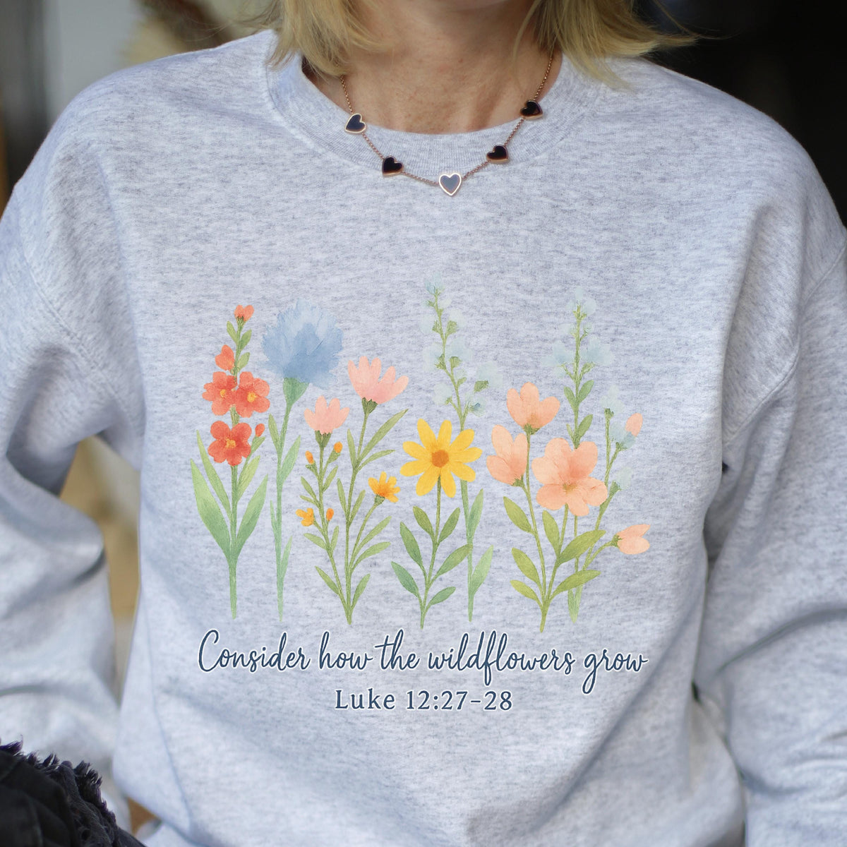 May include: A light gray sweatshirt featuring a watercolor design of colorful wildflowers. The text "Consider how the wildflowers grow" and Bible verse Luke 12:27-28 are printed below the flowers.