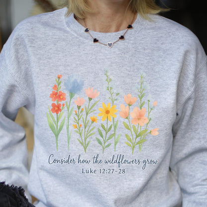 May include: A light gray sweatshirt featuring a watercolor design of colorful wildflowers. The text "Consider how the wildflowers grow" and Bible verse Luke 12:27-28 are printed below the flowers.