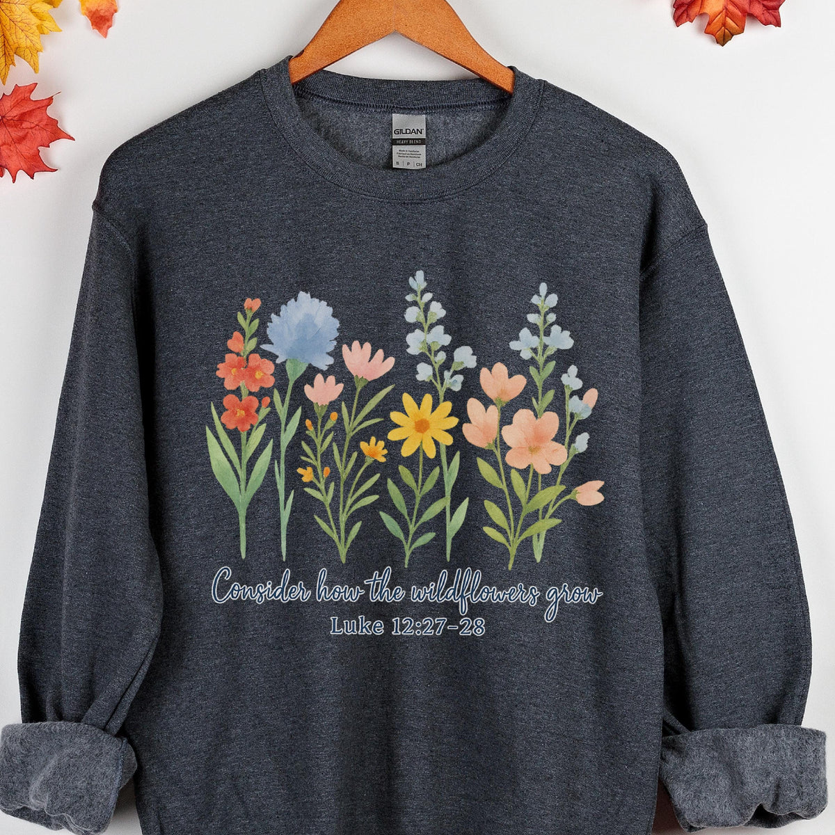 May include: Dark gray sweatshirt featuring a floral design with various colorful wildflowers. The text "Consider how the wildflowers grow" and the Bible verse Luke 12:27-28 are printed below the flowers. The sweatshirt is hanging on a wooden hanger.