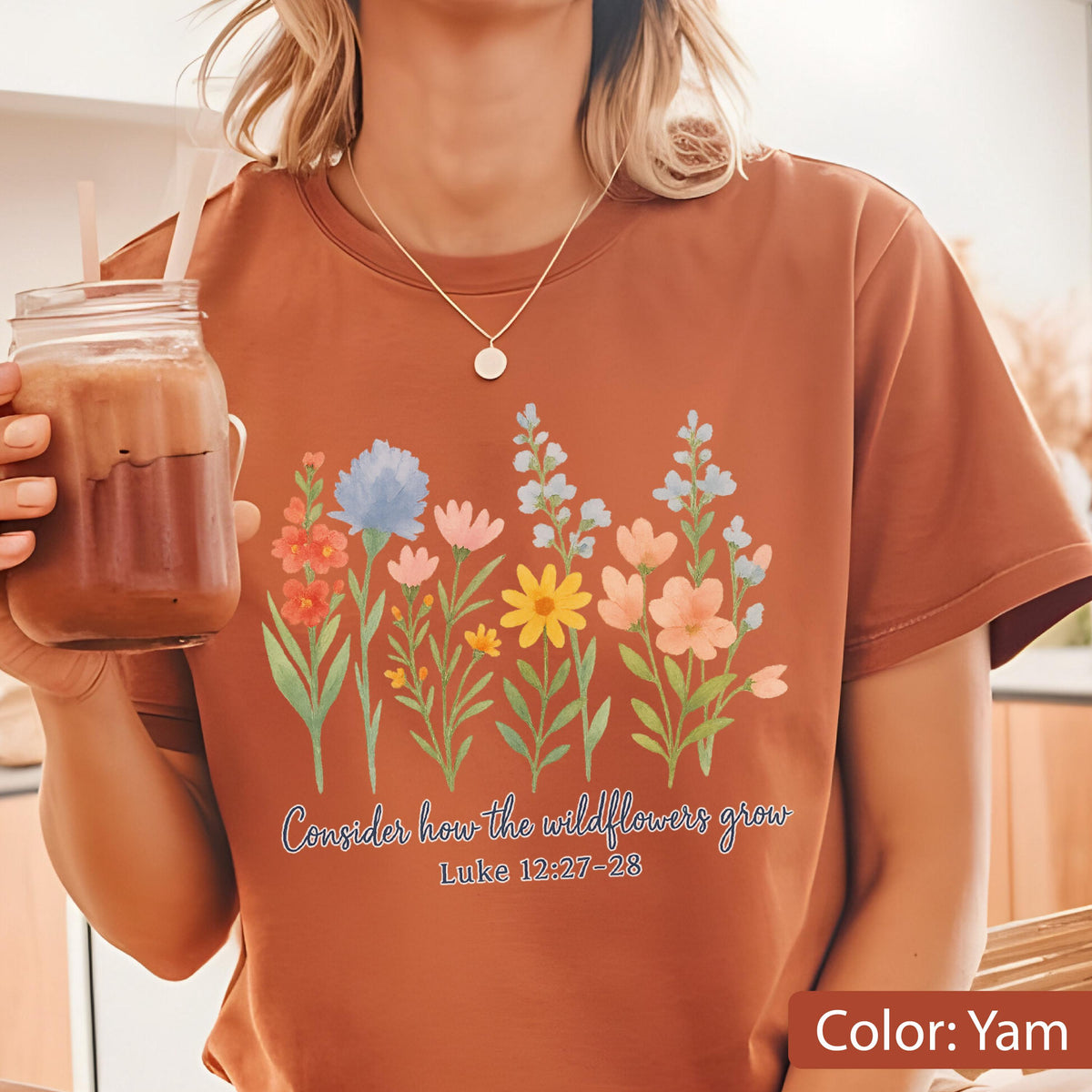 May include: A rust-colored t-shirt featuring a graphic of colorful wildflowers in shades of blue, pink, yellow, and orange. Below the flowers, the text reads "Consider how the wildflowers grow" followed by the scripture reference "Luke 12:27-28".