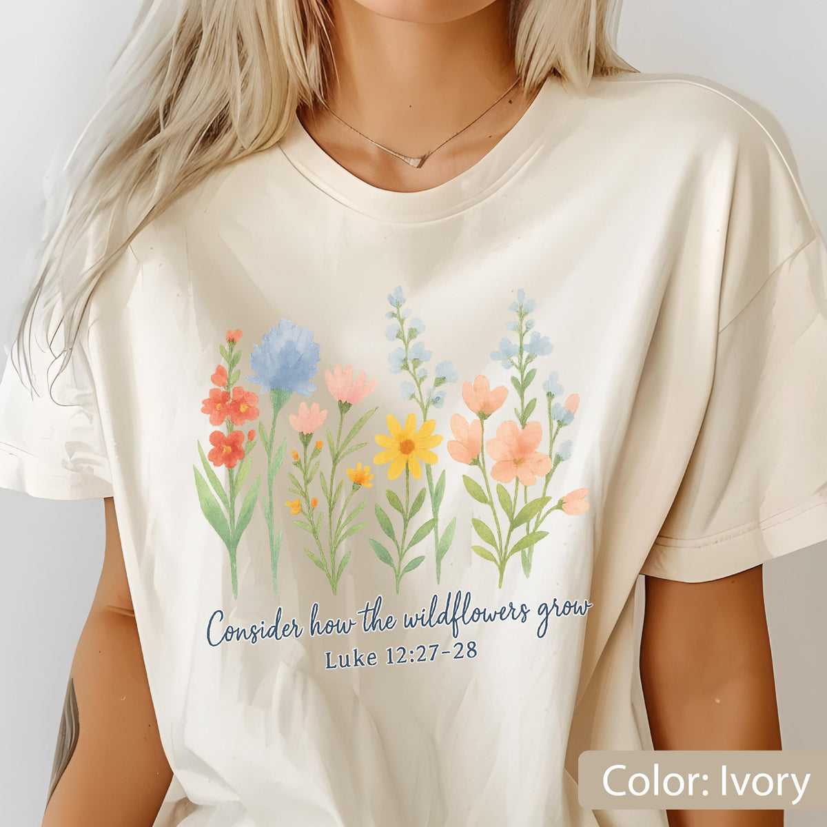 May include: An ivory short-sleeve t-shirt featuring a watercolor-style graphic of various colorful wildflowers in shades of blue, orange, pink, and yellow. Below the flowers, the text reads, "Consider how the wildflowers grow Luke 12:27-28."