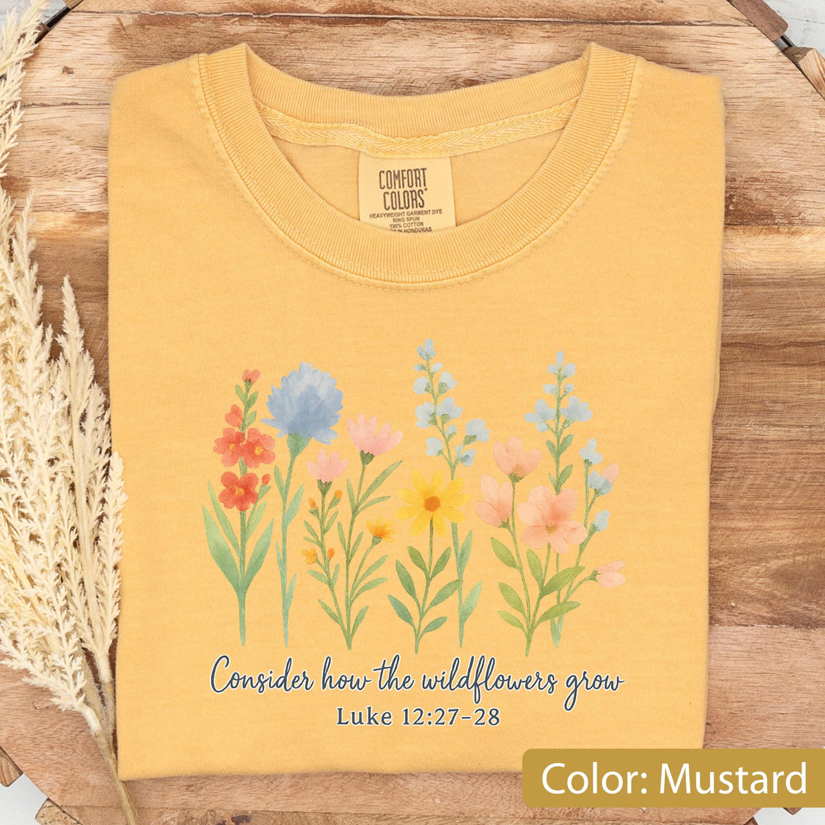 May include: A mustard yellow t-shirt featuring a watercolor-style illustration of various wildflowers in shades of blue, pink, orange, and yellow. Below the flowers, the text reads "Consider how the wildflowers grow" and "Luke 12:27-28".