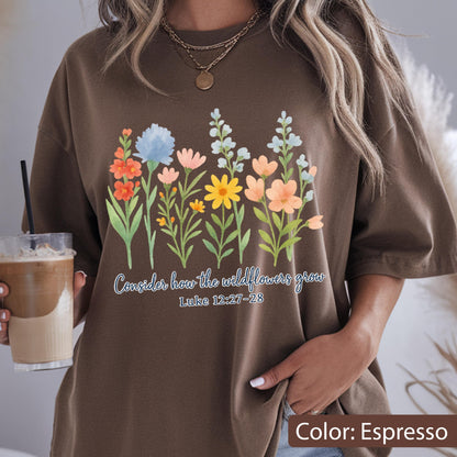 May include: An espresso brown t-shirt featuring a colorful watercolor-style graphic of various wildflowers in shades of orange, blue, pink, and yellow. Below the flowers, the text reads: Consider how the wildflowers grow Luke 12:27-28.