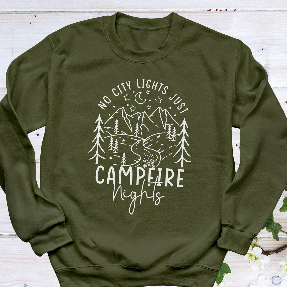 May include: An olive green long-sleeve sweatshirt featuring a white line art graphic of a mountain landscape, pine trees, a crescent moon, stars, and a campfire. The text reads NO CITY LIGHTS JUST CAMPFIRE NIGHTS in a mix of block and script fonts.