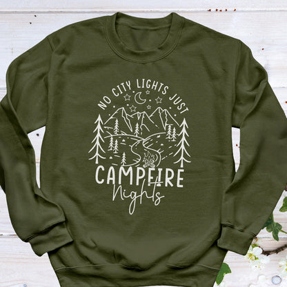 May include: An olive green long-sleeve sweatshirt featuring a white line art graphic of a mountain landscape, pine trees, a crescent moon, stars, and a campfire. The text reads NO CITY LIGHTS JUST CAMPFIRE NIGHTS in a mix of block and script fonts.