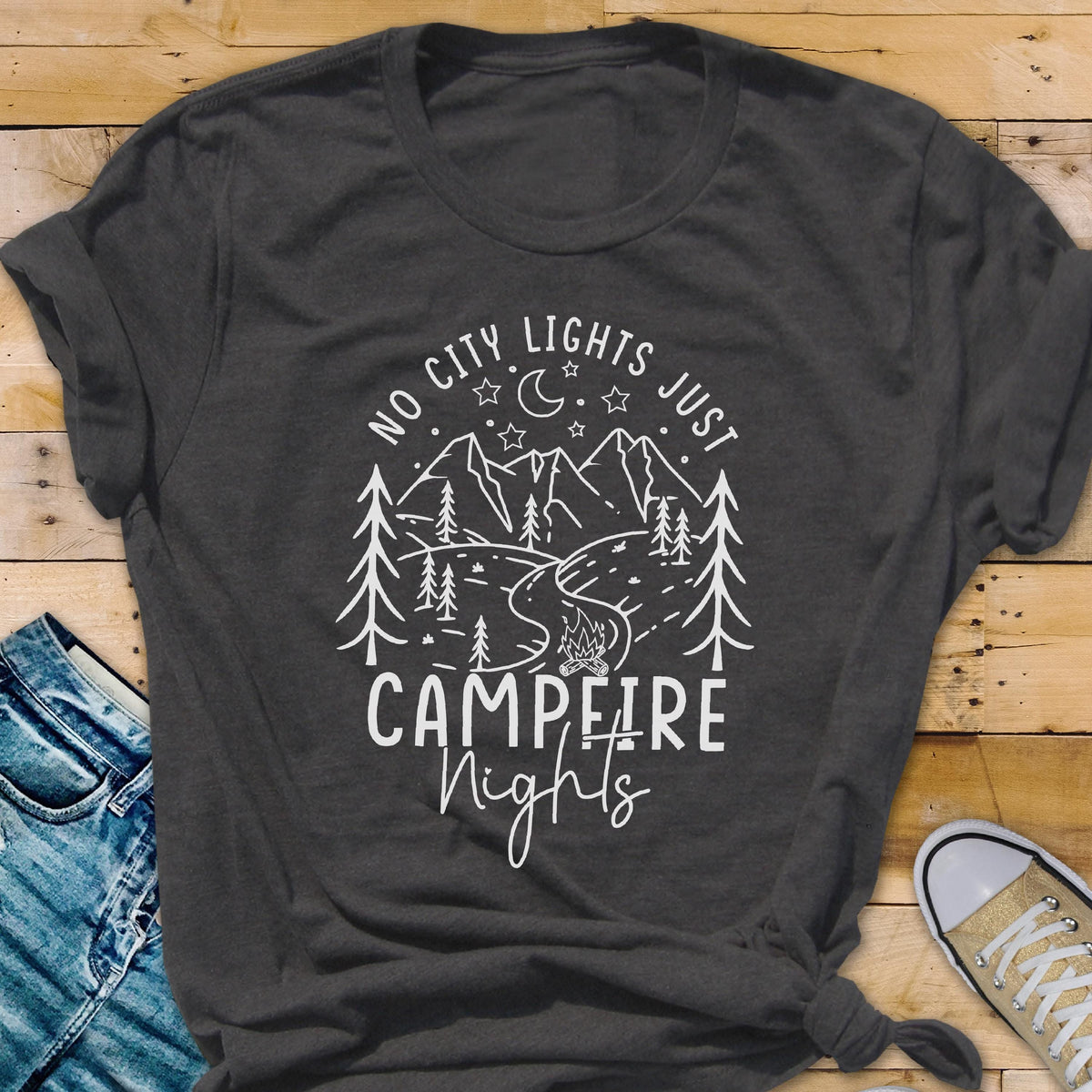 May include: A dark charcoal gray short-sleeved t-shirt featuring a white graphic print of mountains, pine trees, a crescent moon, stars, and a campfire. The text reads NO CITY LIGHTS JUST CAMPFIRE Nights in a mix of block and script lettering.