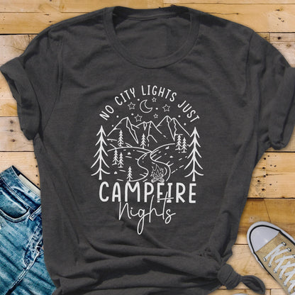 May include: A dark charcoal gray short-sleeved t-shirt featuring a white graphic print of mountains, pine trees, a crescent moon, stars, and a campfire. The text reads NO CITY LIGHTS JUST CAMPFIRE Nights in a mix of block and script lettering.