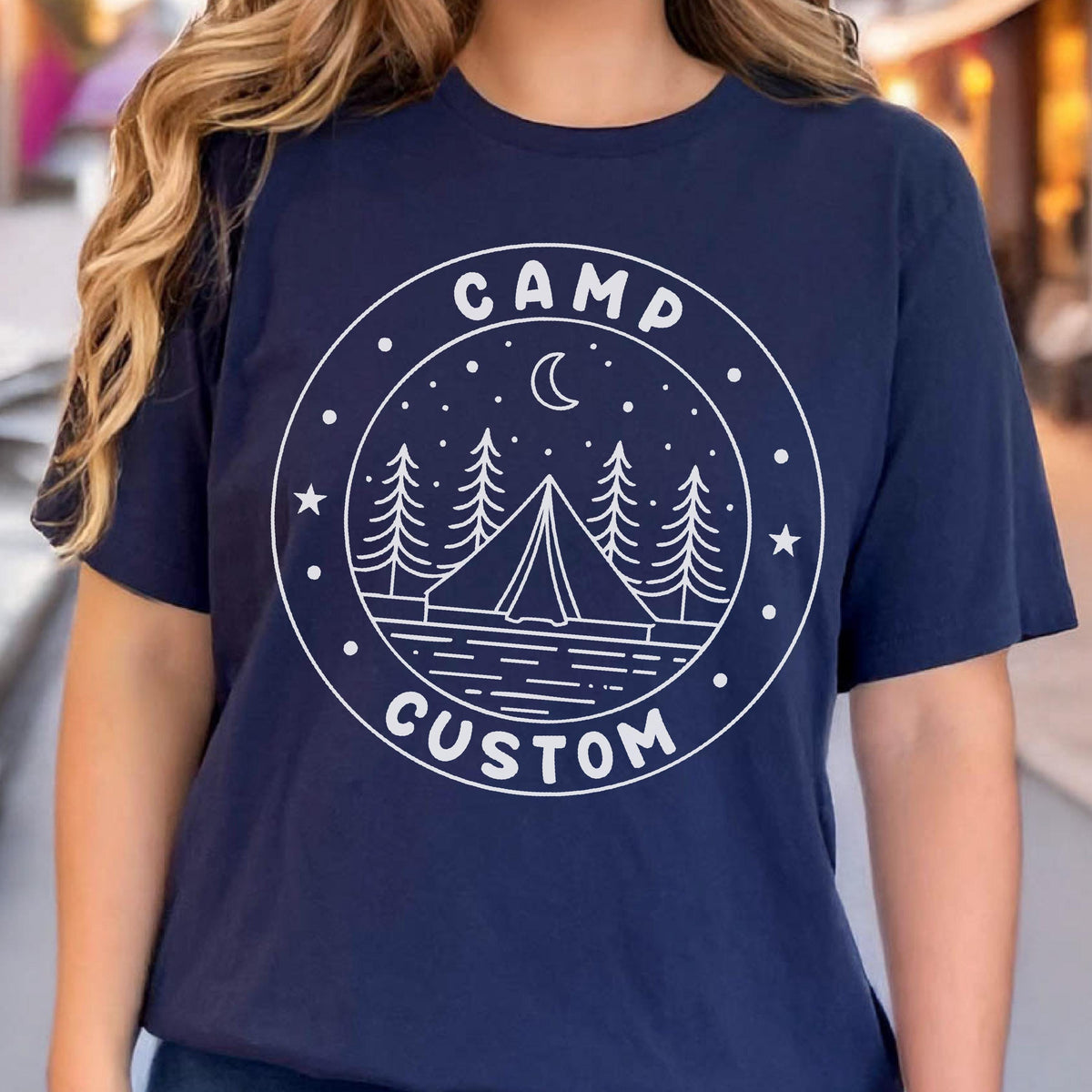 May include: A navy blue short-sleeve t-shirt featuring a white circular graphic on the chest. The design includes a tent, pine trees, a crescent moon, and stars, with the text CAMP at the top and CUSTOM at the bottom of the circular border.