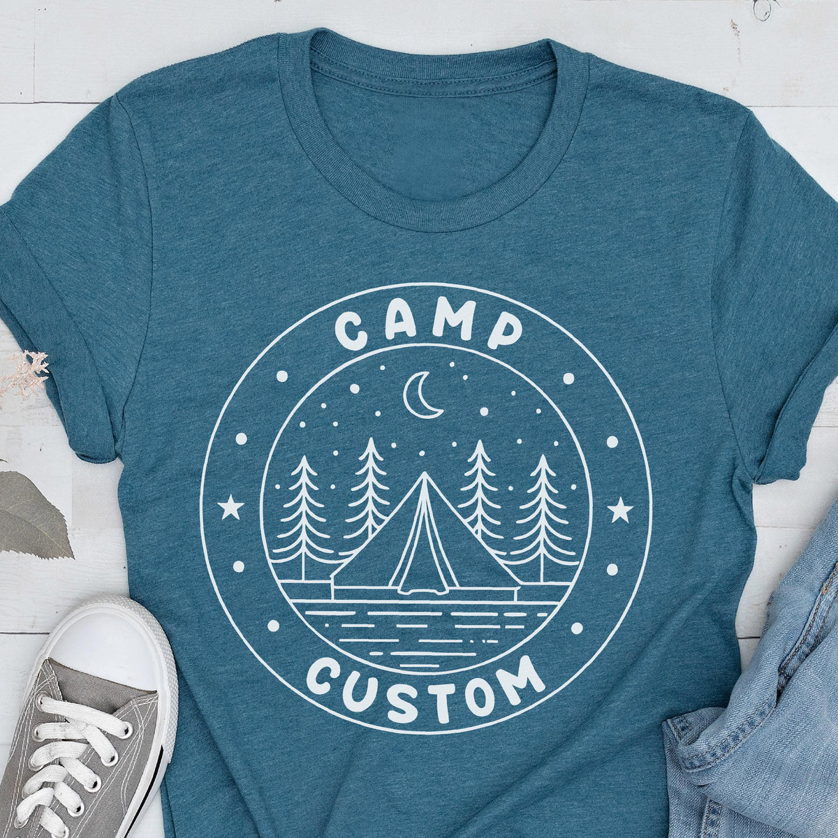 May include: A teal blue short-sleeved t-shirt featuring a white circular graphic print. The design depicts a tent, pine trees, a crescent moon, and stars, with the text "CAMP" at the top and "CUSTOM" at the bottom of the circular border.