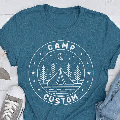 May include: A teal blue short-sleeved t-shirt featuring a white circular graphic print. The design depicts a tent, pine trees, a crescent moon, and stars, with the text "CAMP" at the top and "CUSTOM" at the bottom of the circular border.