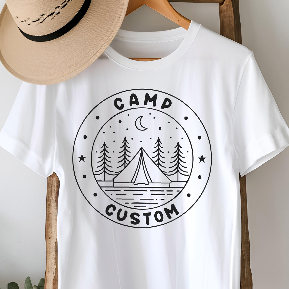 May include: A white short-sleeved t-shirt featuring a black circular graphic on the chest. The design includes a tent, pine trees, a crescent moon, and stars, with the text CAMP CUSTOM printed in a bold, sans-serif font within the circular border.