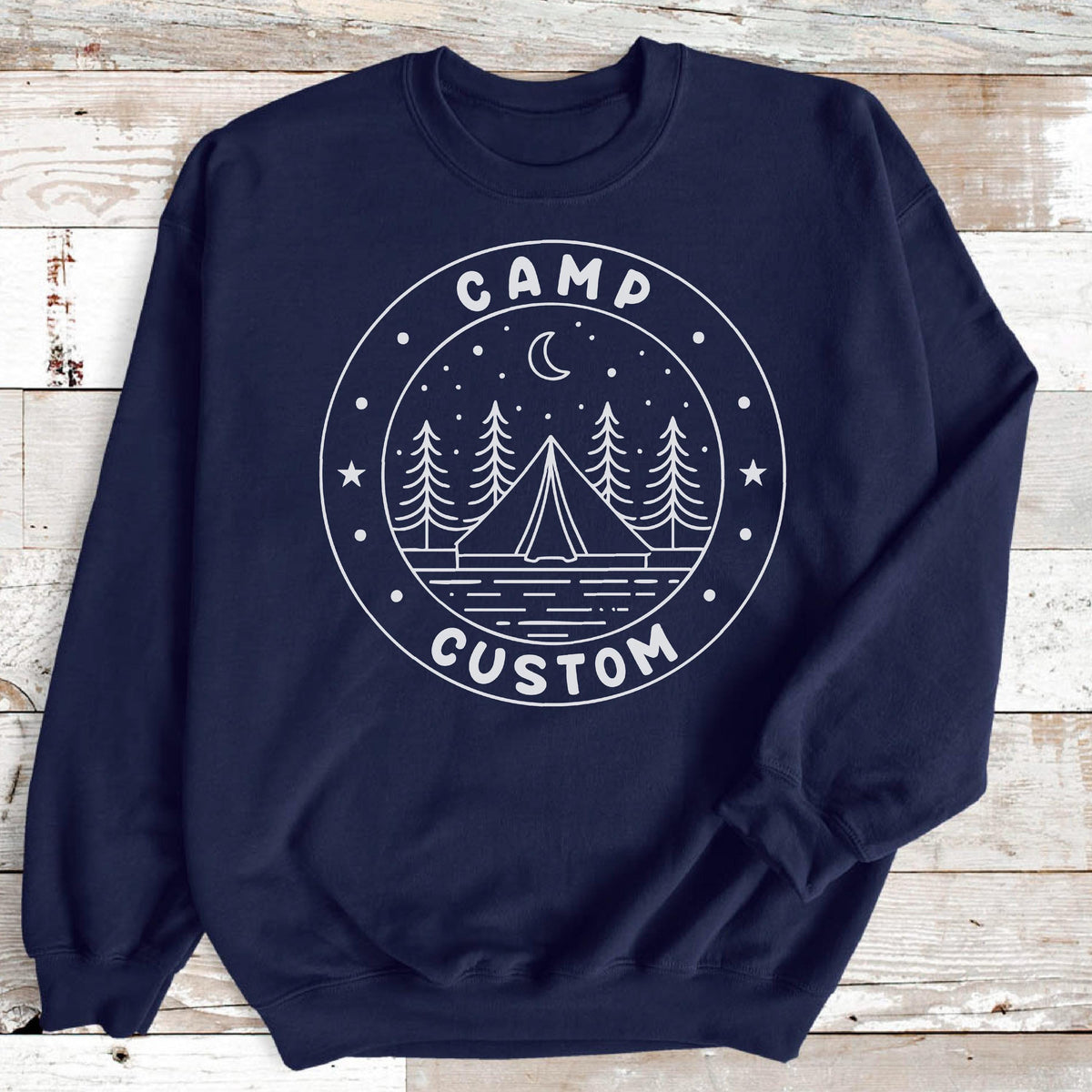 May include: A navy blue long-sleeve sweatshirt featuring a white circular graphic on the chest. The design depicts a tent, pine trees, a crescent moon, and stars, with the text CAMP CUSTOM printed in a circular border around the illustration.